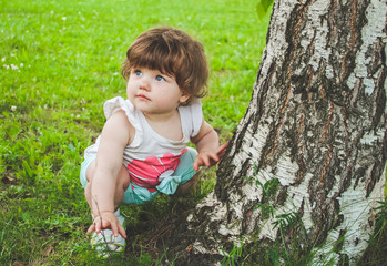 A small child sits on the grass near the tree.