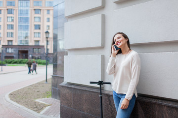 Fototapeta premium Beautiful young girl with long brown hair stopped while riding the scooter, to call a friend on the phone on the background of the gray wall. She is dressed in a white sweater and blue jeans