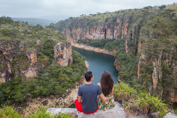 Couple on a rock overlooking a canyon with a river on the bottom and rocky walls covered by green trees. Furnas Canyon is a common tourist destination in Brazil - Powered by Adobe