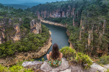 Girl sits on a rock overlooking a canyon with a river on the bottom and rocky walls covered by green trees under a cloudy sky. Furnas Canyon is a common tourist destination in Brazil
