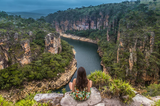 Girl sits on a rock overlooking a canyon with a river on the bottom and rocky walls covered by green trees under a cloudy sky. Furnas Canyon is a common tourist destination in Brazil