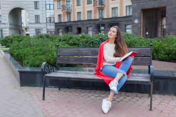 An attractive girl with long brown hair sits on a bench and writes her thoughts on the city background in a red notebook. She wears a white sweater, blue jeans and a red plaid.