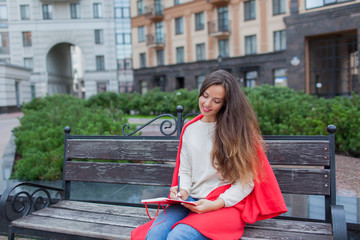 An attractive girl with long brown hair sits on a bench and writes her thoughts on the city background in a red notebook. She wears a white sweater, blue jeans and a red plaid.