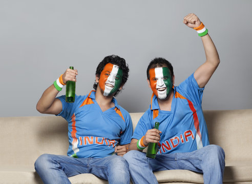 Male cricket fans with painted face in Indian tricolor enjoying game while having drink at home 