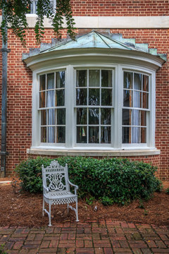 Curved exterior bay window with a metal roof is part of an southern mansion. A white iron chair sits in the yard in front of the bay window.