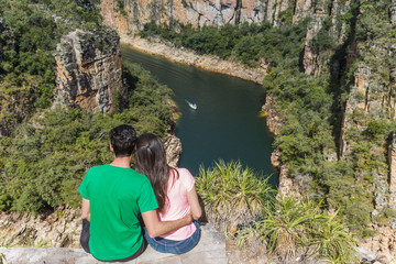 Couple on a rock overlooking a canyon with a river on the bottom and rocky walls covered by green trees. Furnas Canyon is a common tourist destination in Brazil