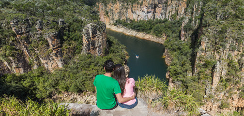 Couple on a rock overlooking a canyon with a river on the bottom and rocky walls covered by green trees. Furnas Canyon is a common tourist destination in Brazil