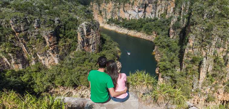 Couple on a rock overlooking a canyon with a river on the bottom and rocky walls covered by green trees. Furnas Canyon is a common tourist destination in Brazil