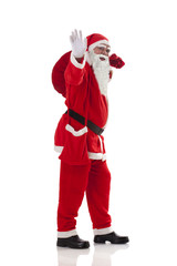 Full length of Santa Claus waving while carrying sack full of presents over white background 