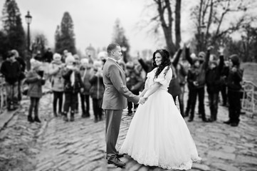 Gorgeous wedding couple holding hands on the pavement while crowd of kids takes pictures of them. Black and white photo.