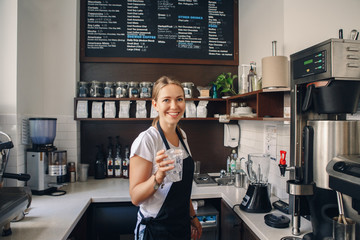 Portrait of  happy beautiful young Caucasian smiling woman barista holding plastic cup with ice. Server making cold drink in coffee shop. Toned with film filters. - obrazy, fototapety, plakaty
