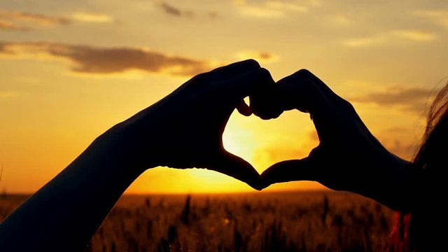 Girl forns a heart with her hands over the sun at sunset in a field of wheat.
