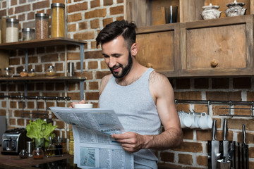 handsome smiling bearded man in pajamas drinking coffee and reading newspaper at home