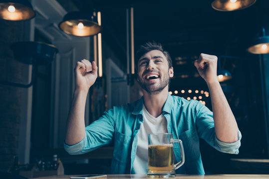 Goal! Cheerful young man is happy because of the goal of his favourite team. He is watching game at the fancy cafe with a glass of beer, in casual jeans outfit in pub