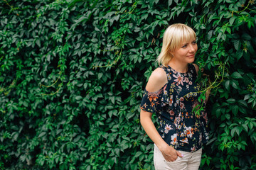 Cute young blonde stylish woman in pretty blouse in flowers over green grape leaves background. Beautiful smiling girl