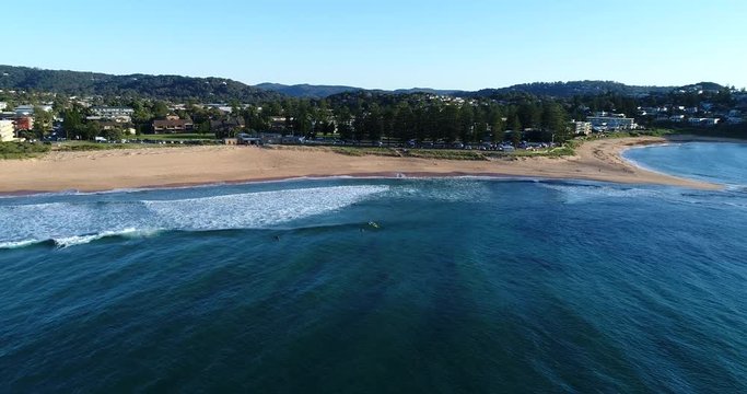 Aerial elevated photo along waterline on Mona Vale Narrabeen beach with surfers, clean sand and blue waves going towards the coast and recreation park. 