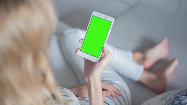 Young Woman in white jeans laying on couch holds SmartPhone with pre-keyed green screen. Perfect for screen compositing. Made from 14bit RAW. 10bit ProRes 444