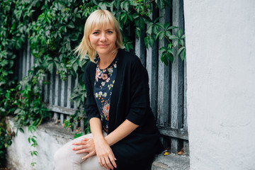 Cute young blonde beautiful stylish woman in pretty blouse in flowers and black cardigan sitting on fence over green grape leaves background.