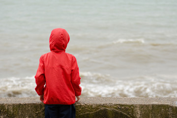 Boy in red jacket with hood standing back at the pier on the background of sea waves