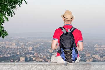 Young man in backpack and hat sitting on background of large city below. Back view