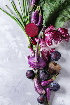 Assortment raw organic of purple vegetables mini eggplants, spring onion, beetroot, radicchio salad, plums, kohlrabi over gray concrete background. Top view with space