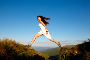 Young woman in white dress running in the mountains. Woman jumping on mountain peak rock. Beautiful girl looking happy and smiling