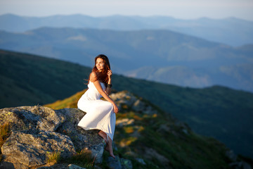 Naklejka na ściany i meble Young woman sitting on a rock. Hair blowing in the wind. Beautiful woman in a long white dress in the mountains