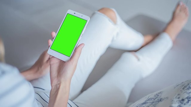 Young Woman in white jeans sitting on couch holds mobile phone with pre-keyed green screen. Perfect for screen compositing. Made from 14bit RAW. 10bit ProRes 444