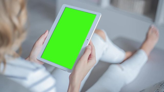 Young Woman in white jeans sitting on couch holds Tablet PC with pre-keyed green screen. Perfect for screen compositing. Made from 14bit RAW. 10bit ProRes 444