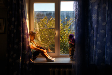 Cute female child with blonde hair sitting on windowsill, looking out window at sunset.