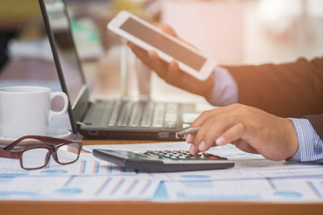 A businessman analyzing investment charts at his workplace and using his tablet and touch calculator.