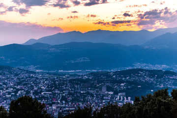 Aerial view of the lake Lugano surrounded by mountains and evening city Lugano on during dramatic sunset, Switzerland, Alps. Travel