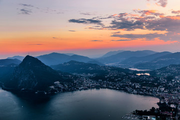 Aerial view of the lake Lugano surrounded by mountains and evening city Lugano on during dramatic sunset, Switzerland, Alps. Travel