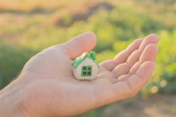 Woman's hand holds small plasticine toy house on the background of ground and green grass. Buying a house concept.