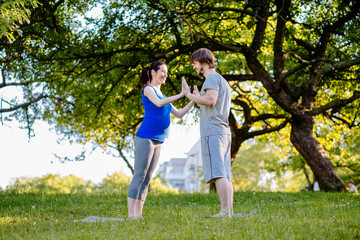 Naklejka na ściany i meble Pregnant couple workout outdoor - restraining palm to palm while standing on yoga mat in park. Parenthood, sport and healthy life concept.