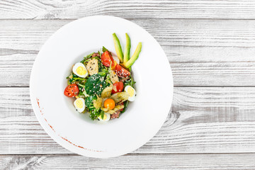 Fresh salad with tuna, spinach, arugula, codfish, artichokes, quail eggs, avocado, cherry tomatoes, physalis and sesame on wooden background close up. Mediterranean food