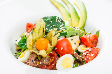 Fresh salad with tuna, spinach, arugula, codfish, artichokes, quail eggs, avocado, cherry tomatoes, physalis and sesame on wooden background close up. Mediterranean food