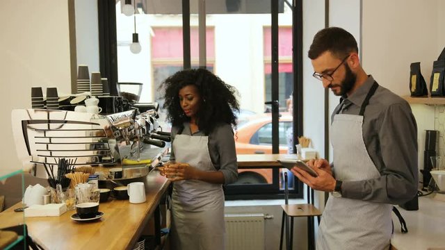 Africana female and caucasian male barista working in modern coffee cafe, woman making coffee for the customer, man using laptop for checking the order. View from the side.