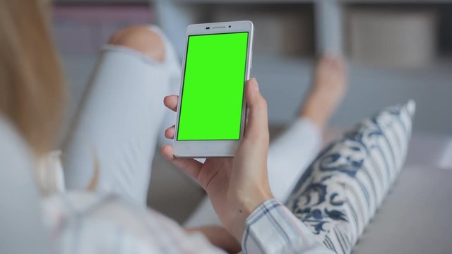 Young Woman in white jeans laying on couch holds mobile phone with pre-keyed green screen. Perfect for screen compositing. Made from 14bit RAW. 10bit ProRes 444