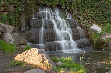 Small waterfall in Alexandria park, Belaya Tserkov, Ukraine. Shot on a sunny hot summer day. Golden setting sun light on the rock near the river with the waterfall in the background