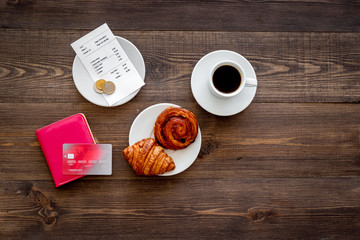 Pay bill at cafe by card. Purse, bill and bank card near coffee and croissant on dark wooden table top view copyspace