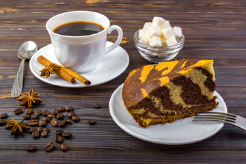 A chocolate cake on a white plate with a fork and a cup of black tea. A tea spoon. anise, coffee beans and a bowl of sugar cubes on dark wooden background