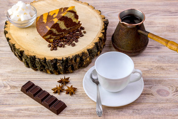 Obraz na płótnie Canvas A white coffee cup with a chocolate cake on wooden stump, tea spoon, coffee beans, anise, chocolate bar and bowl with sugar cubes on a bright wooden table