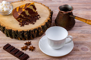 A coffee cup with a piece of cake on wooden stump, a chocolate bar, anise, coffee beans and bowl with sugar cubes on a wooden table