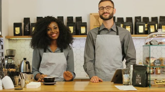 Portrait of happy caucasian man and african woman standing behind light wooden counter, crossing hands and smiling on camera in the cafe.