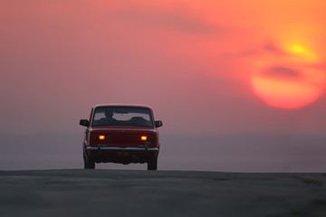 Fototapeta na wymiar Old red car driving on the road towards sunrise