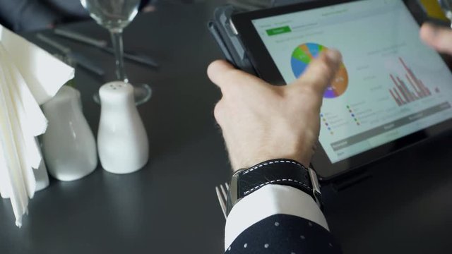 Close up hands of worker discussing business with tablet in cafe during business lunch. Closeup shooting of arms moving pictures on screen, graphics. Male arm wearing classic suit, watches holding