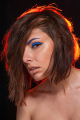 Unretouched portrait of a young pretty caucasian brunette girl posing in a studio on black background with bright orange backlight. She has messy hair and bright blue eye liner and beige lip gloss.