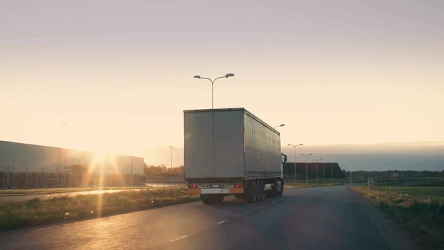 Follow-up Shot of a Semi-Truck with Cargo Trailer Moving on a Highway. White Truck Drives Through Industrial Warehouse Area on an Empty Road with Sun Shining in the Background. 4K UHD.