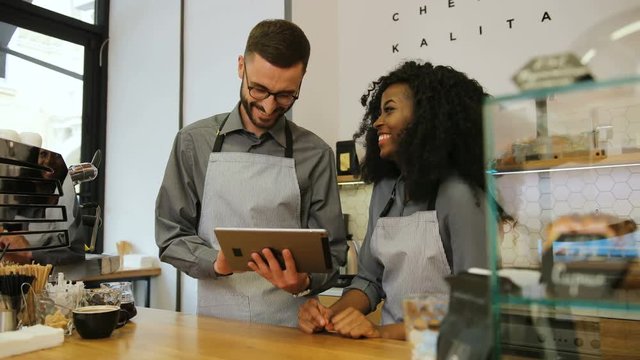 Two young barista man and woman using tablet computer during a break on the work in the coffee shop. Man barista showing funny videos to his coworker.